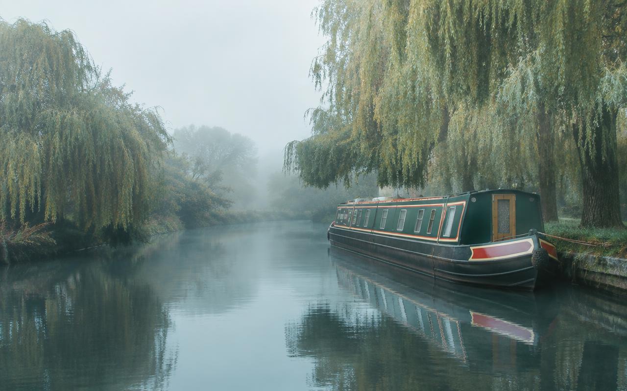 Green narrowboat moored on a misty English canal under willow trees
